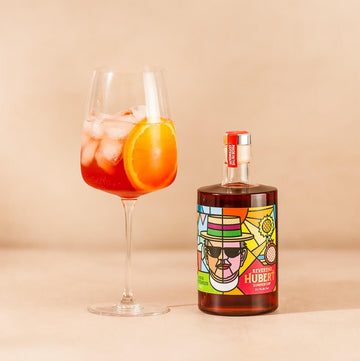 A bottle of Reverend Hubert Summer Cup with a Cotswold Spritz cocktail next to it, the cocktail is in a large wine glass with lots of ice and an orange slice