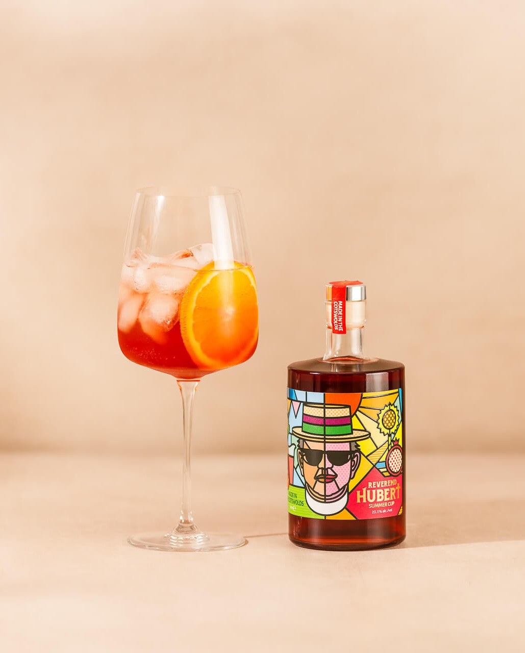 Bottle of Reverend Hubert Summer Cup with a cocktail called The Reverend's Cotswold Sprtiz - a cocktail in a large wine glass with ice and a slice of orange